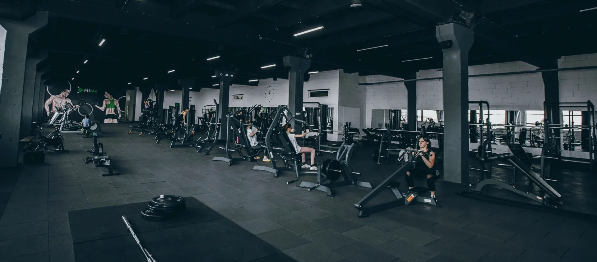 Інтер'єр залу FitMe — тренажери, настінний мурал FitMe і високі вікна у нічному грейді